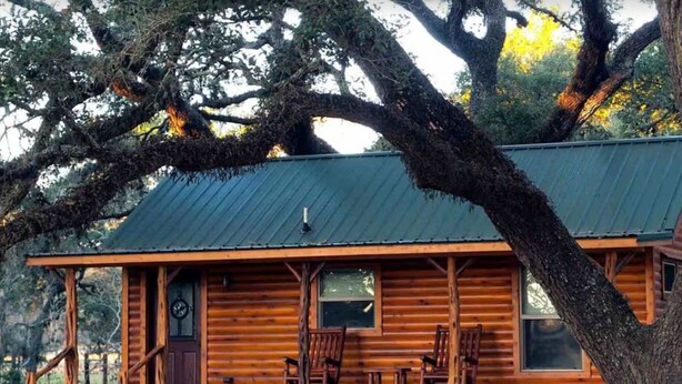 Une cabane rustique en rondins de bois, dotée d’un porche, de fondations en pierre et de grandes fenêtres.