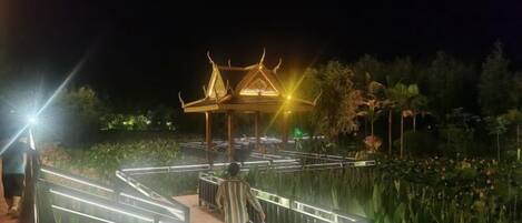 A night-time view of a traditional Thai pavilion with a tiled roof, surrounded by a wooden deck and lush greenery.
