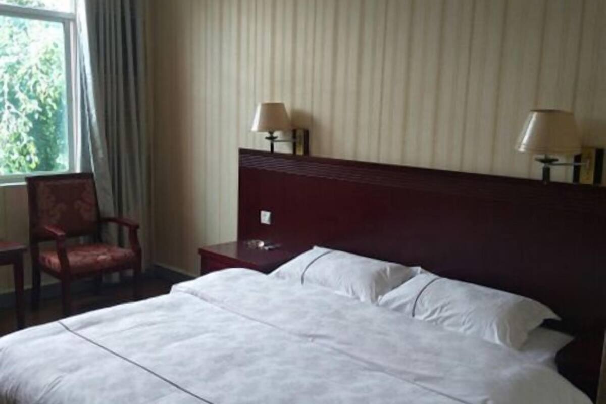 A hotel room with a large bed, two bedside lamps, a wooden headboard, a chair, and a window with curtains.