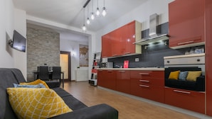 A modern kitchen with red cabinets, a black countertop, and a built-in oven. There is a dining area with a table and chairs, and a living room with a sofa and cushions. - Turin World Apartment by Wonderful Italy (Turin)