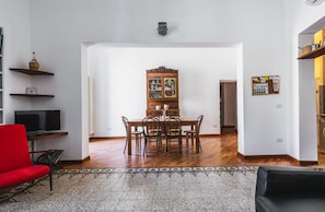 Un comedor con una mesa y sillas de madera, una silla roja, un televisor sobre un mueble bajo y una alfombra estampada.