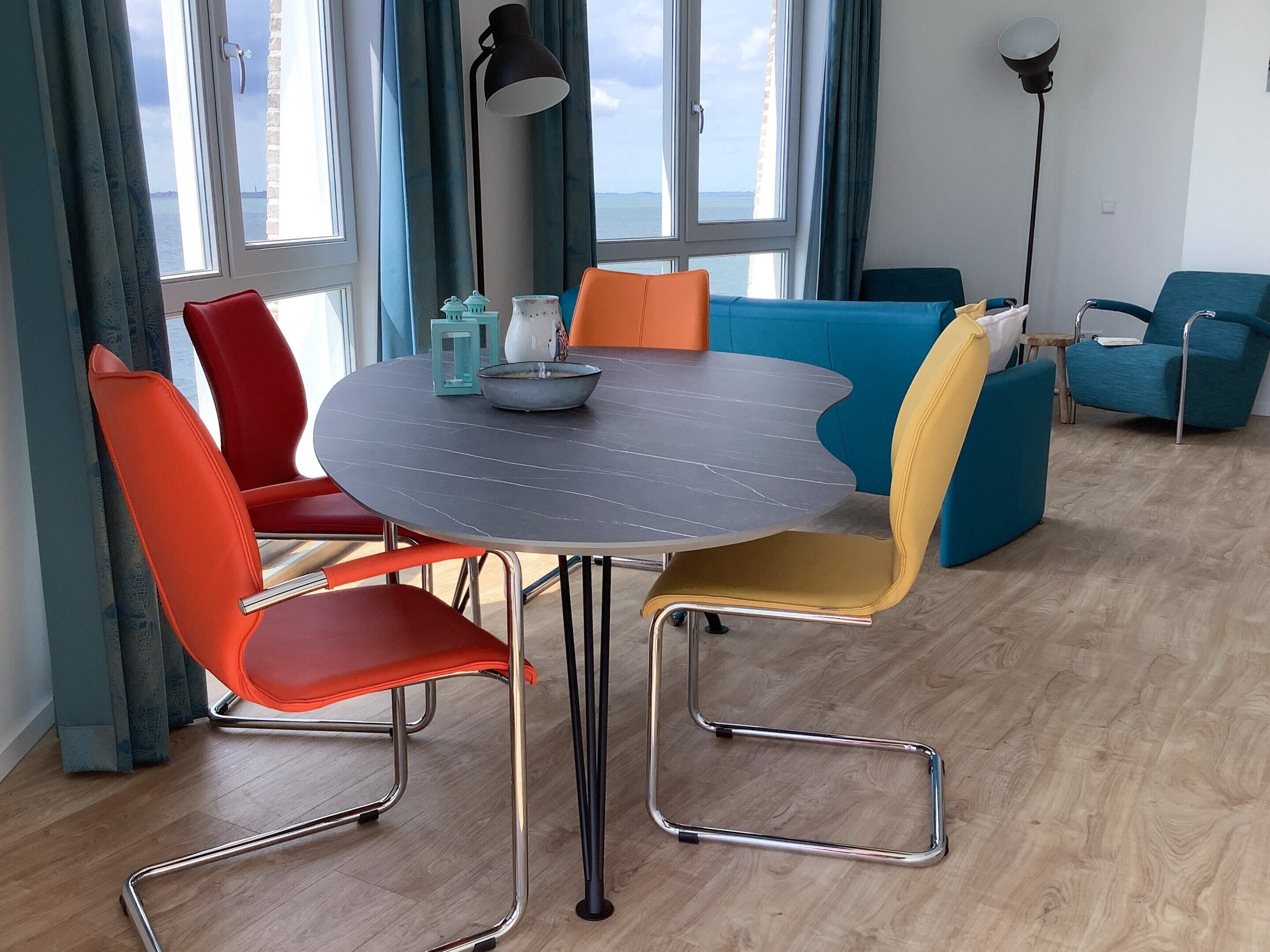 Ein modernes Wohnzimmer mit einem runden Esstisch, bunten Stühlen und großen Fenstern mit Blick auf das Meer.
