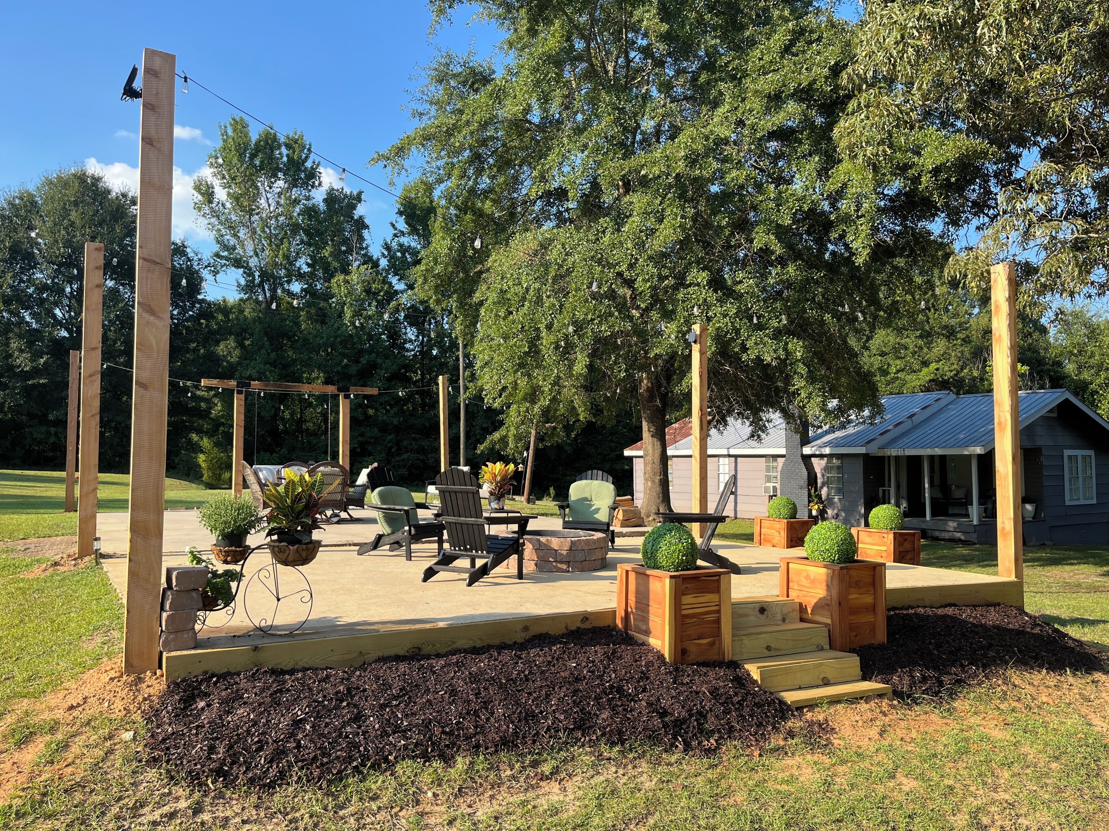An outdoor seating area with wooden chairs and tables, surrounded by trees and a house in the background.