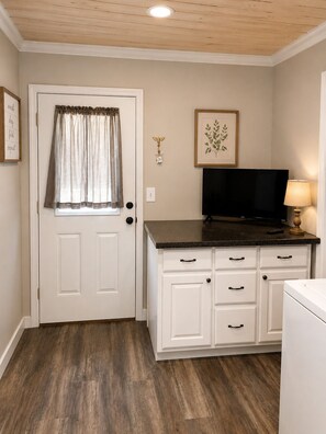 A room with a white door, a window with a curtain, a flat-screen TV on a black stand, a white cabinet with drawers, and a wooden floor.