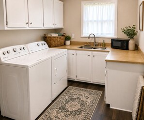 A laundry area with a front-loading washer and dryer, a sink, a microwave, and a window with sheer curtains.