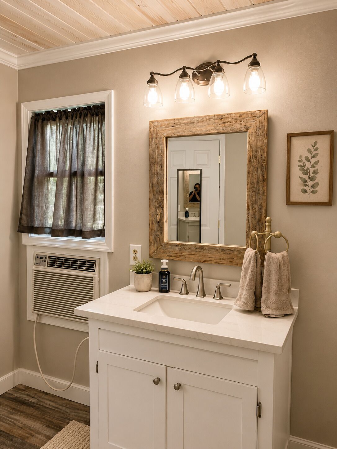 A bathroom with a white vanity, a mirror with a wooden frame, a window with curtains, a wall-mounted light fixture, and a framed plant.