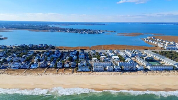 An aerial view of a coastal resort with multiple blue-roofed buildings, a sandy beach, and a large body of water. - Sunshine and Bethany Beach Dreams (Bethany Beach)