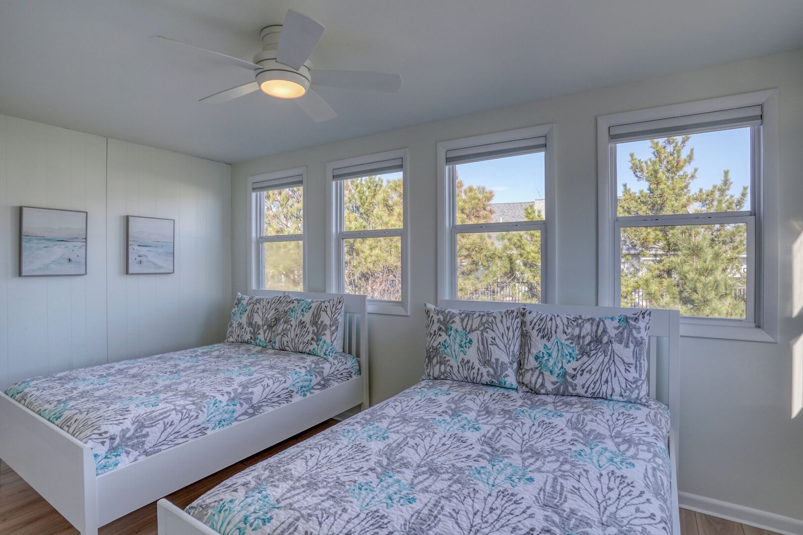 Un dormitorio con dos camas individuales, ventilador de techo y amplios ventanales con vista a árboles.