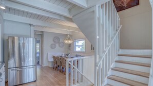 A spacious kitchen with a stainless steel refrigerator, a dining area with wicker chairs, and a staircase with white railings. - Sunshine and Bethany Beach Dreams (Bethany Beach)
