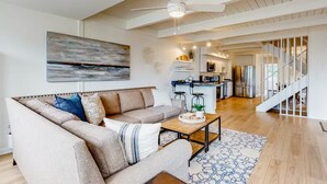 A spacious living room with a large sofa, a coffee table, and a ceiling fan. A kitchen area is visible in the background with a staircase leading to an upper level. - Sunshine and Bethany Beach Dreams (Bethany Beach)