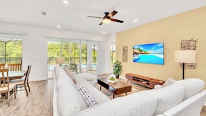 A spacious living room with a large white sofa, a wooden coffee table, and a television mounted on the wall.