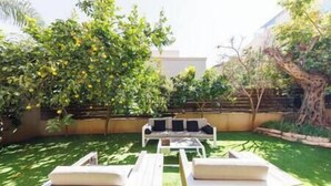A patio with a white table and two white chairs, surrounded by green grass and trees. - Five Stars Villa09 (Hod Hasharon)