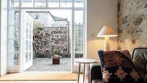 A cozy living room with a fireplace, a comfortable sofa, and a view of a stone wall and garden through large windows. - Hideout by Stay Lagom (Ballycastle)