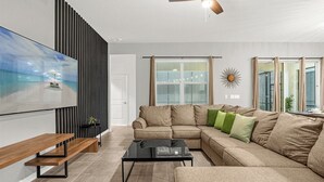 A modern living room with a sectional sofa, a flat-screen TV, a ceiling fan, and a large window with curtains.