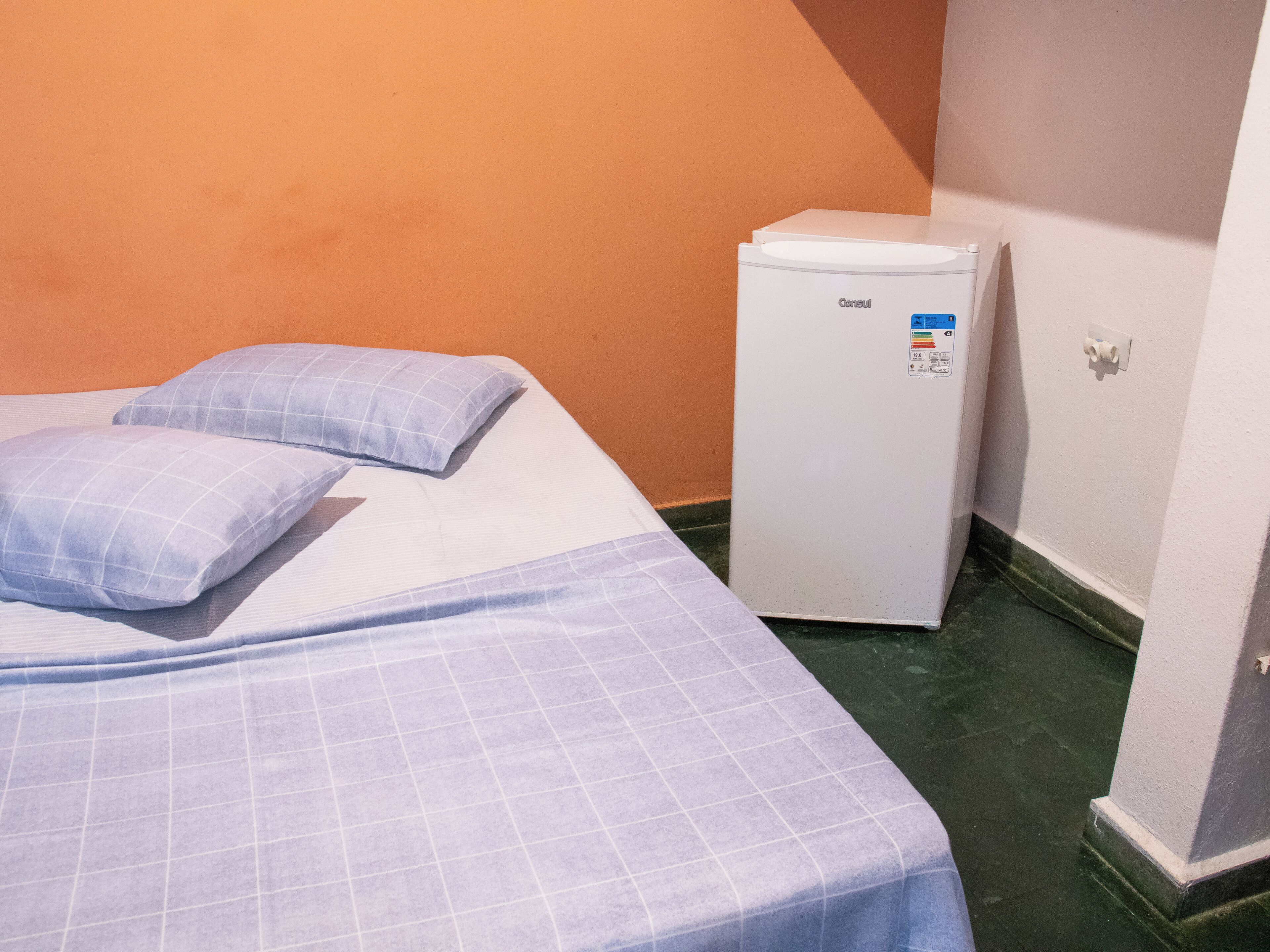 A single bed with a blue checkered bedspread and pillows, a white bedside table, and a white refrigerator in a room with orange walls and a green floor.