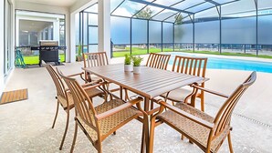 Une terrasse extérieure avec une table en bois et des chaises en osier, un barbecue, et une vue sur une piscine et une pelouse verdoyante.