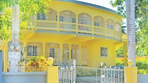 Un edificio amarillo de dos pisos con balcones, una entrada con reja y un pequeño jardín.