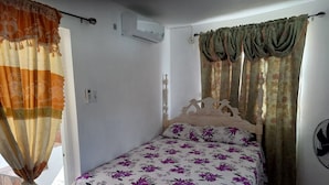 Un dormitorio con cama, cortinas, aire acondicionado, una mesita de noche y un ventilador de pared.