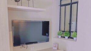 Habitación con televisión montada en la pared, un escritorio pequeño con una silla, un radiador y una ventana con plantas en macetas.