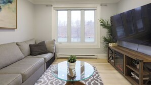 Una sala de estar con un sofá, una mesa de centro de vidrio, un televisor sobre un mueble de madera y un ventanal con vistas.