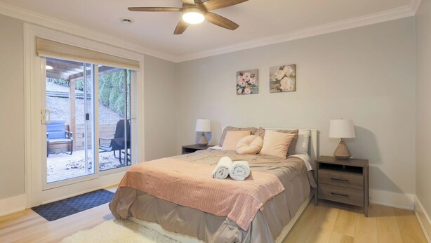 Un dormitorio con una cama, dos mesitas de noche con lámparas, un ventilador de techo, un ventanal corredero a un espacio exterior y dos cuadros de flores colgados en la pared.