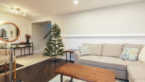 Una sala moderna con un sofá color beige, una mesa de centro de madera, una mesa de vidrio con base de metal, un pequeño árbol de Navidad y un espejo en la pared.