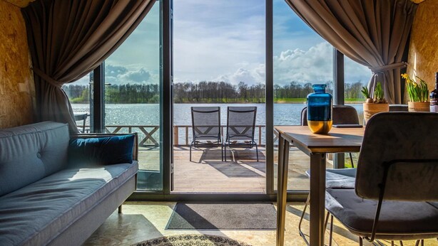 A cozy room with a large window, a sofa, a table with chairs, and a view of a lake and forest. - Modern Chalet 5 km From Kampen (Kampen)