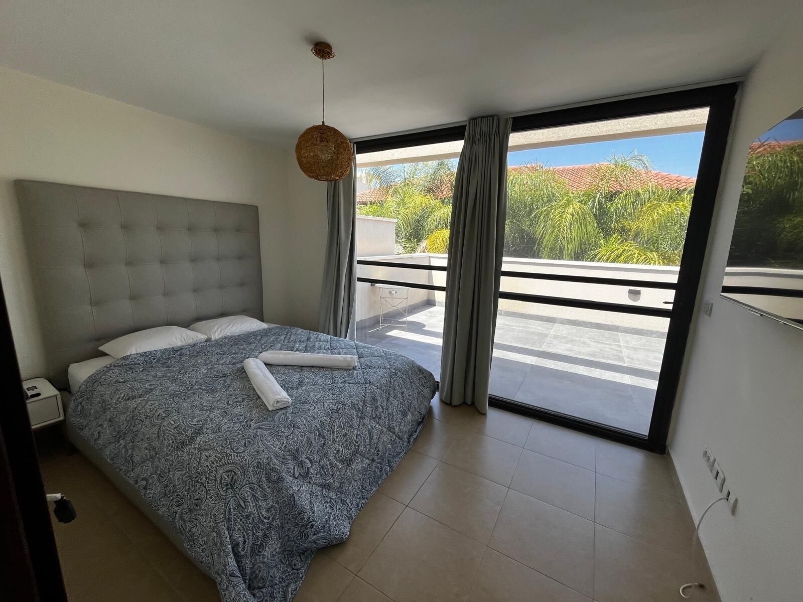 Ein modernes Schlafzimmer mit einem großen Bett, einer hängenden Leuchte und Blick auf einen Balkon mit Grünpflanzen.