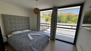 Ein modernes Schlafzimmer mit einem großen Bett, einer hängenden Leuchte und Blick auf einen Balkon mit Grünpflanzen.