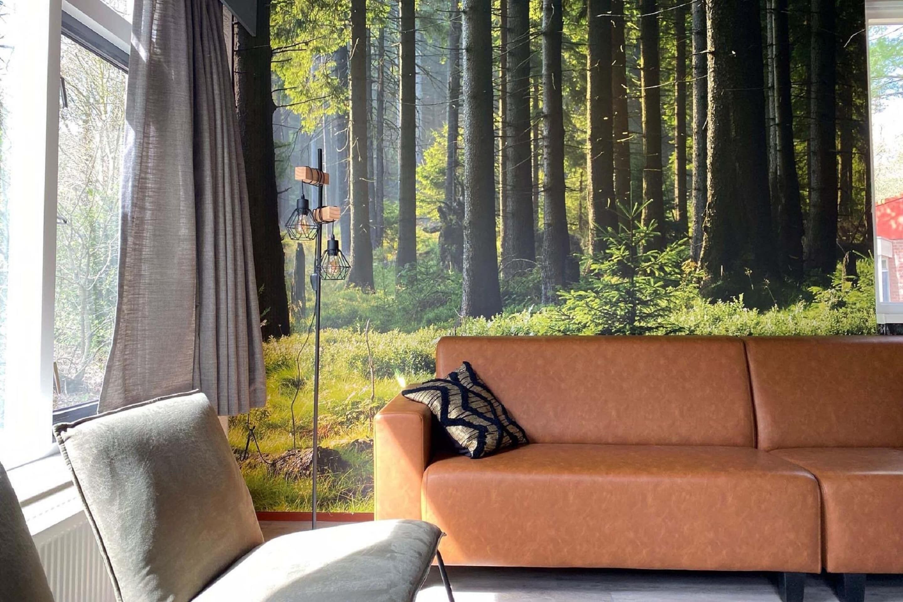 A room with a large window, a brown sofa, a chair, and a forest-themed wall mural.