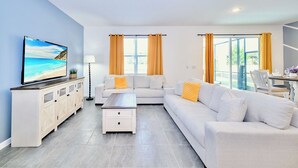 A modern living room with a flat-screen TV, white sofas, a coffee table, and large windows with curtains.