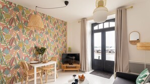 Un salon avec une table blanche, deux chaises, un canapé, un téléviseur et un mur décoré d’un motif.