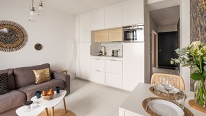 A modern living room with a sofa, coffee table, and dining area. It includes a kitchen with white cabinets and a built-in oven. - Ty Peskaiden 1 -  à Cancale, vue mer (Cancale)