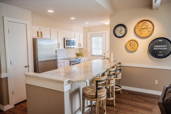 A kitchen with a central island, bar stools, a refrigerator, microwave, and wine-themed wall decor.