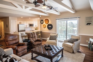 A spacious living room with leather chairs, a tufted ottoman, and a view of the kitchen and balcony.