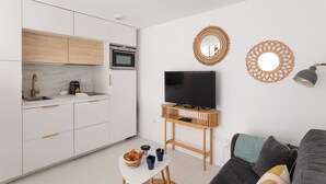 Et moderne køkken med hvide skabe, indbygget ovn og kaffemaskine. Der er et fjernsyn på et træstel, et vægtæppe og en lampe. En sofa med puder er i opholdsstuen.