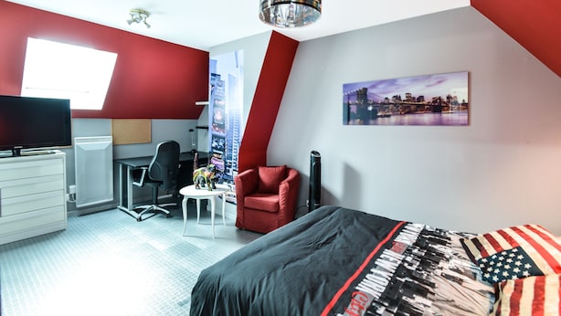 Ett modernt sovrum med en röd accentvägg, en målning som föreställer en stadsbild, en säng med ett svart och rött överkast, ett skrivbord med en stol och en tv.