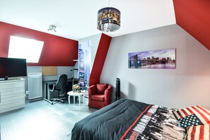 Quarto moderno com parede de destaque vermelha, pintura de paisagem urbana, cama com cobertura preta e vermelha, escrivaninha com cadeira e televisão.