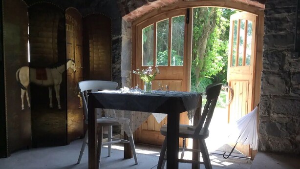 A rustic stone room with a wooden table and chairs, a horse mural, and a large window with a view of greenery.