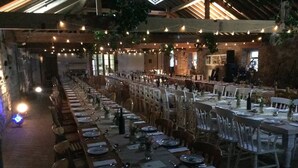 A large, rustic dining hall with long tables set for a meal, string lights overhead, and a stone wall in the background.