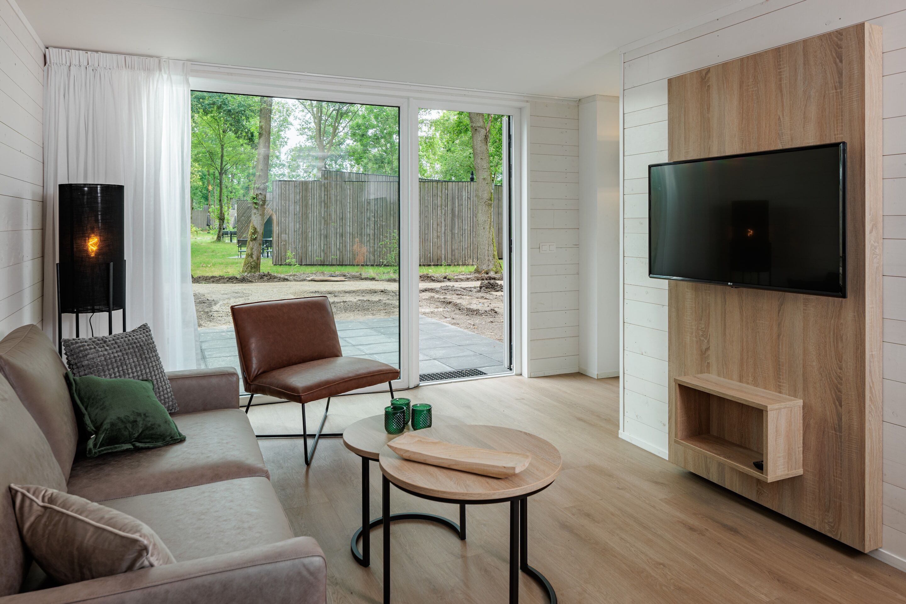 A modern living room with a sofa, a chair, a coffee table, a TV mounted on a wooden wall, and a sliding glass door leading to an outdoor area.