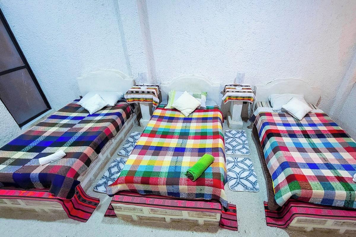Habitación con tres camas, cada una con una colcha de cuadros coloridos. Hay almohadas y una manta en cada cama. El suelo es de baldosas y hay una ventana en el lado izquierdo.