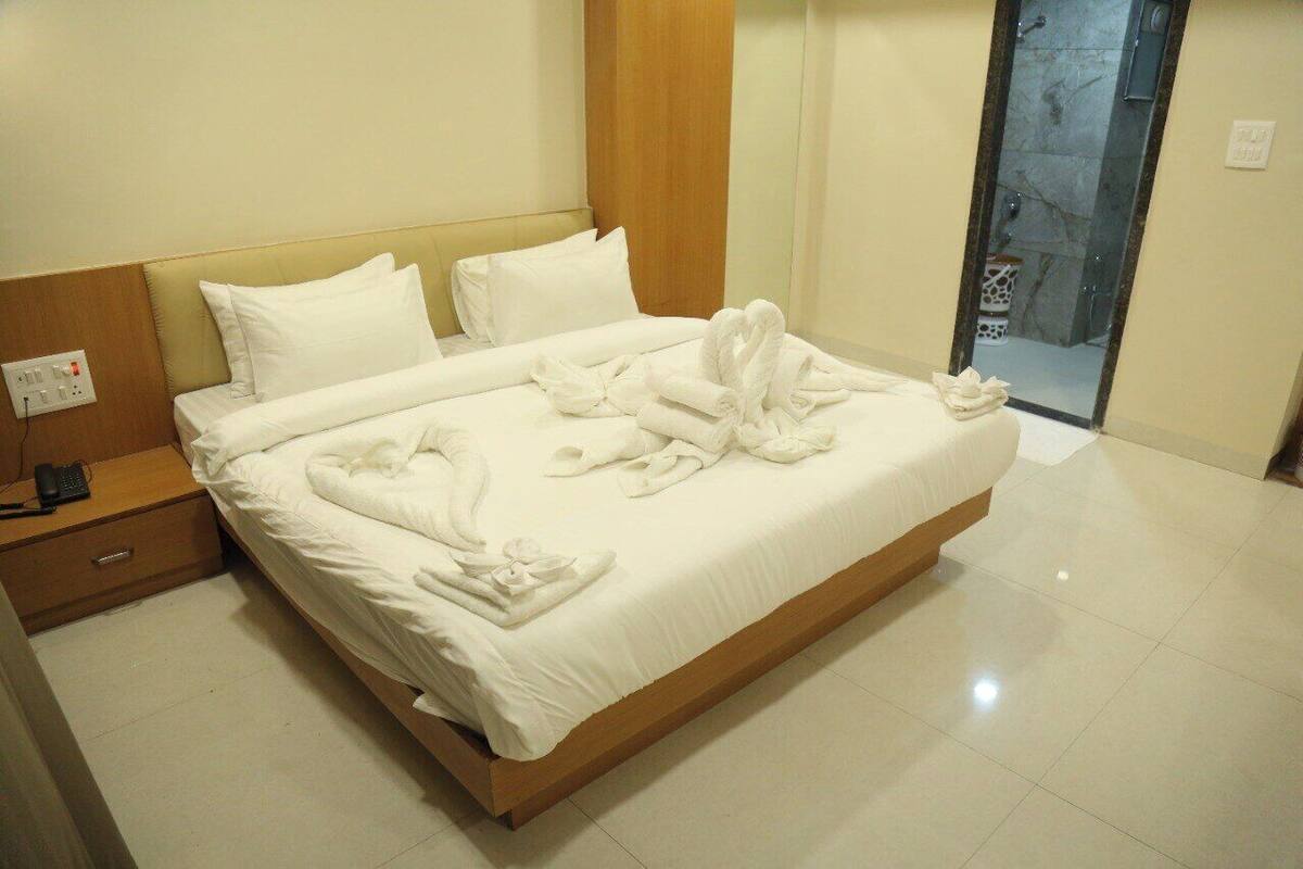 A neatly made bed with white linens and towels arranged on it, a wooden nightstand with a phone, and a bathroom visible through a glass door.