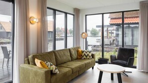 Una sala de estar moderna con un sofá verde, una mesa redonda y grandes ventanas que ofrecen vistas a un cuerpo de agua y edificios.