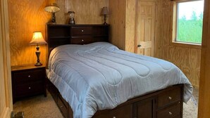 Een slaapkamer met een houten plafond, een bed met een blauwe quilt, twee bedlampen en een raam met uitzicht op groen.