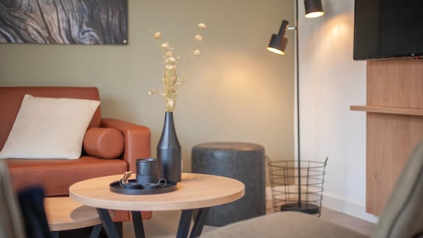 A modern living room with a sofa, a round table, a vase with dried flowers, a floor lamp, and a flat-screen TV mounted on the wall.