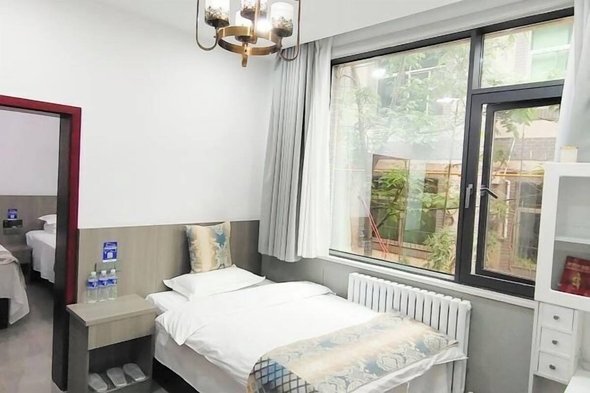 A hotel room with a bed, a nightstand, a window with a view of greenery, a radiator, and a chandelier.