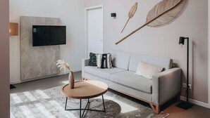 A modern living room with a grey sofa, a round wooden coffee table, a mounted flat-screen TV, and decorative wall art. - Private Indoor Pool Home in Verviers (Breskens)
