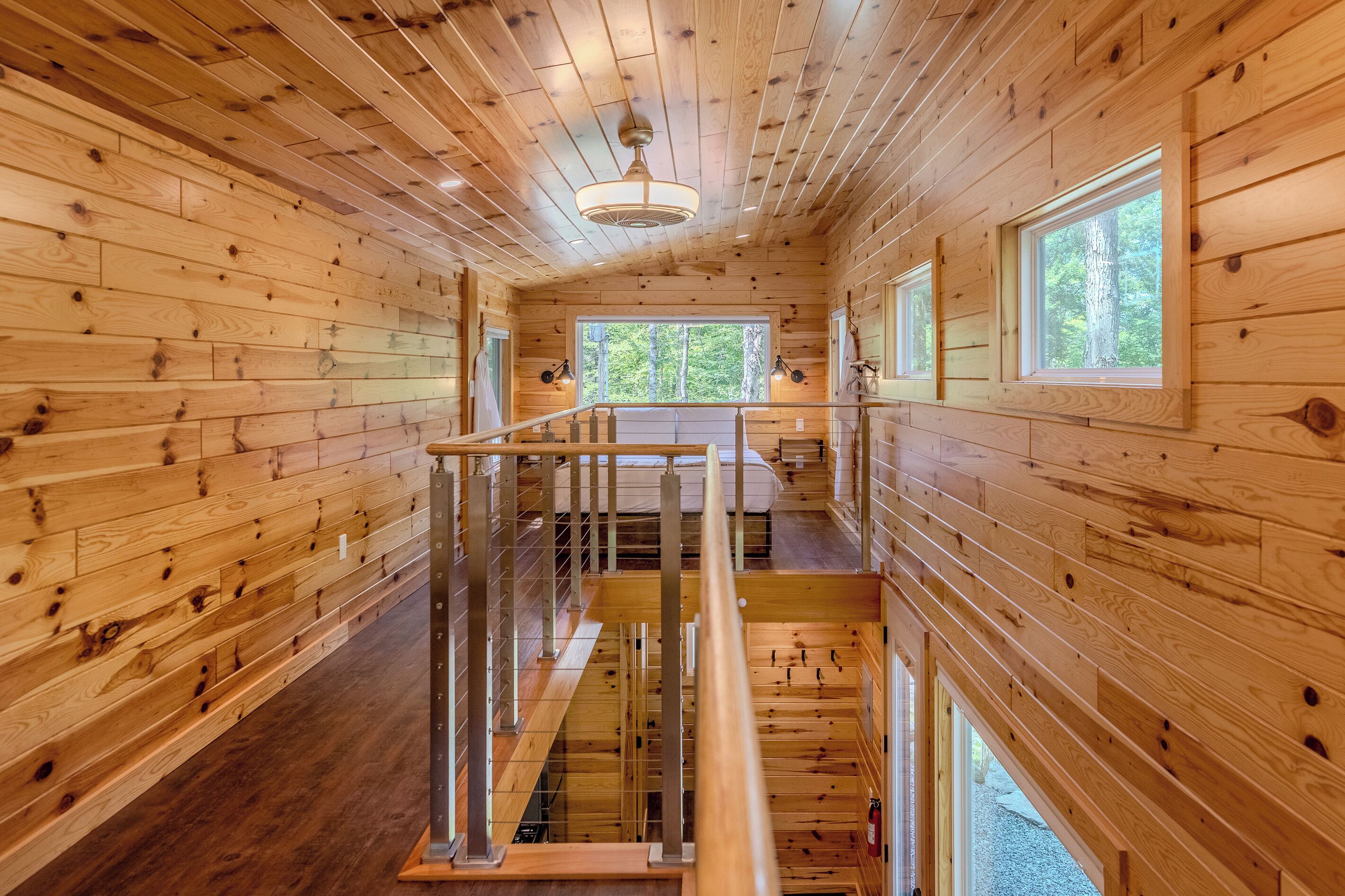 A wooden cabin interior with a central staircase, wooden walls, and a window providing a view of the outdoors.
