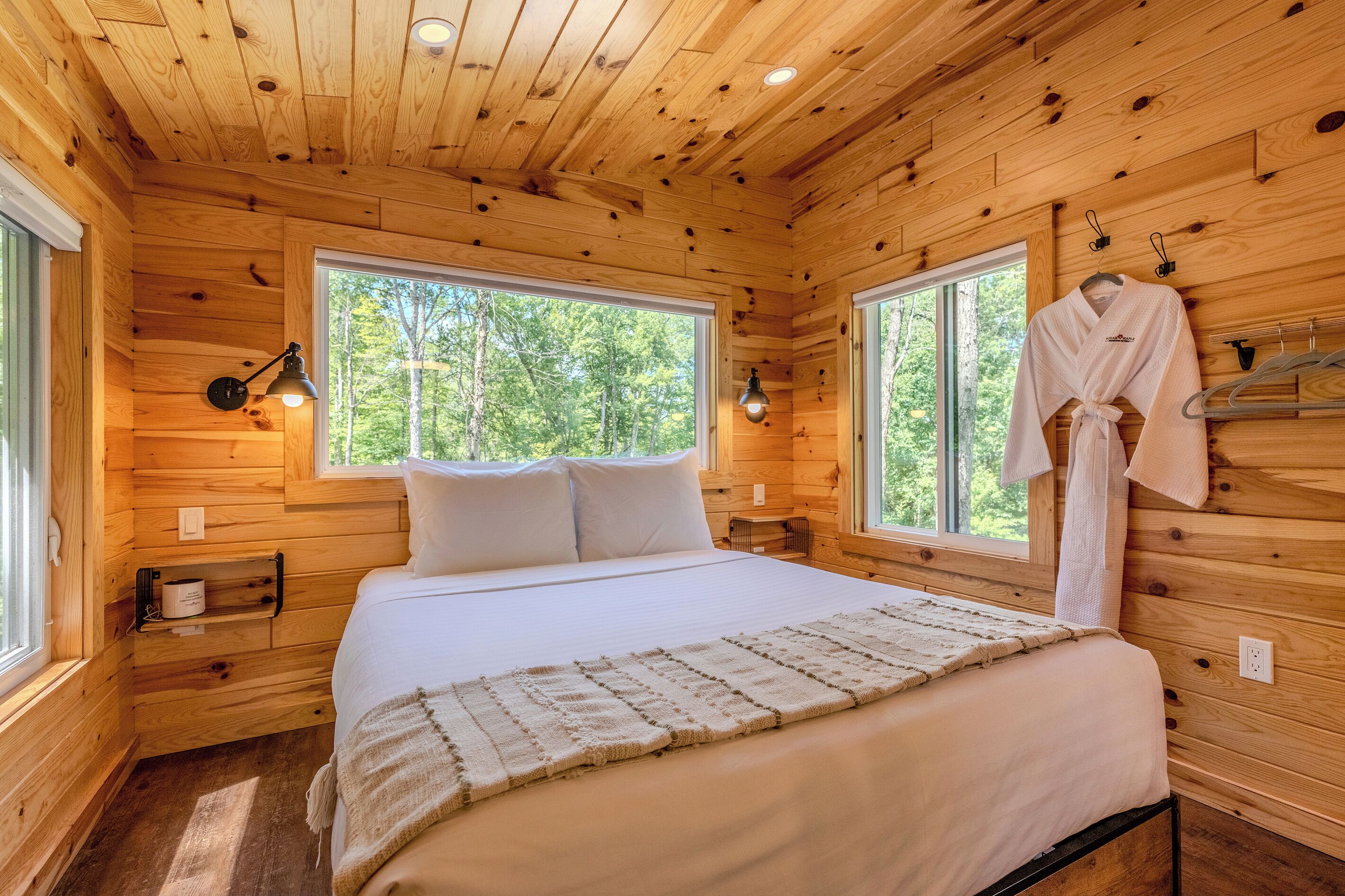 Ein Schlafzimmer einer Holzhütte mit einem Bett, zwei Fenstern, einem aufgehängten Bademantel und einer Wandlampe.